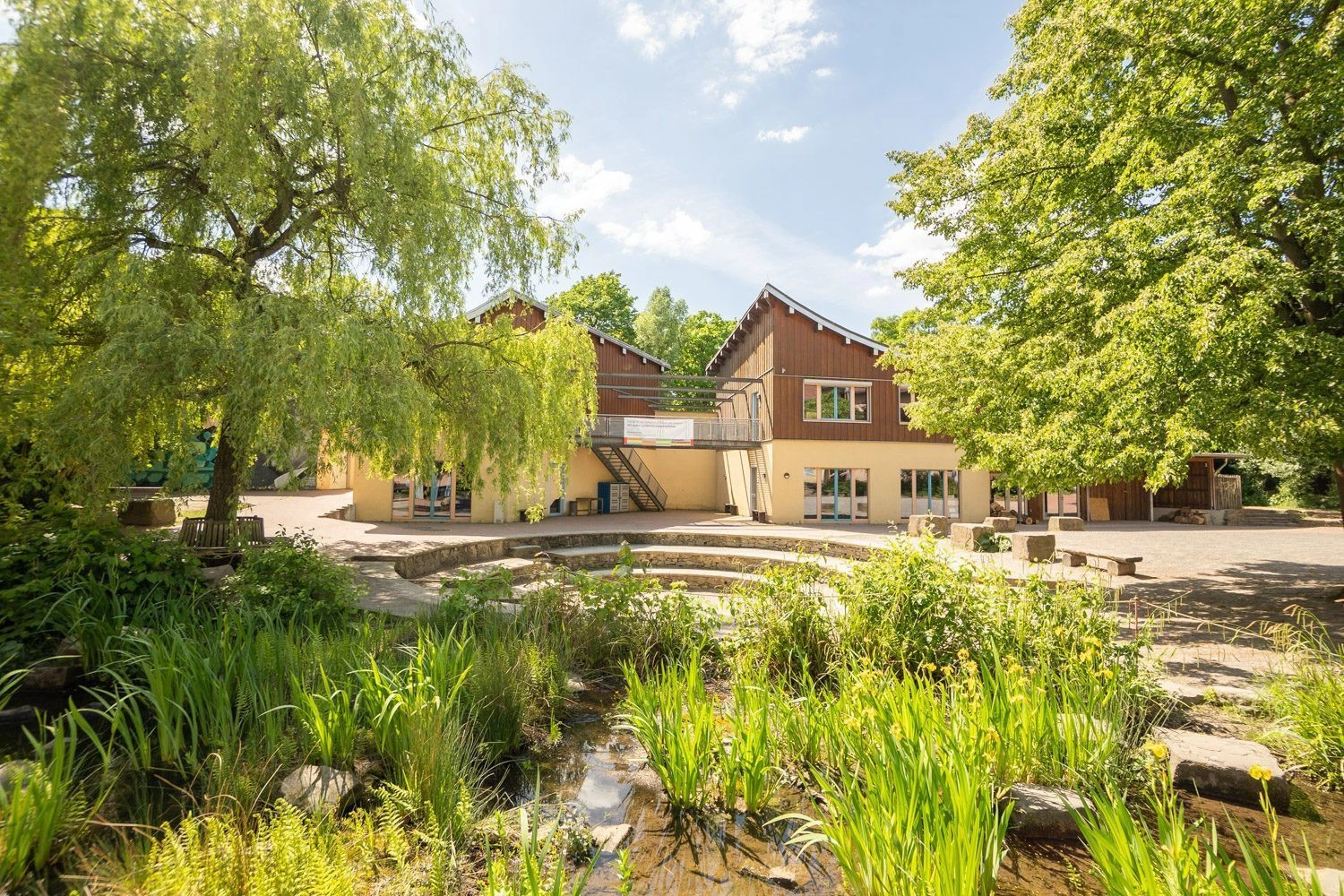 Ansicht auf ein Gebäude der Schule, mit einer Wasserfläche und einer Arena im Vordergrund.